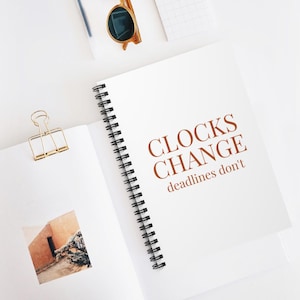 Pode incluir: Um caderno de espiral branco com o texto castanho CLOCKS CHANGE deadlines don't na capa. Um clipe de metal dourado está preso à página e uma pequena fotografia quadrada de um edifício é visível na página aberta.