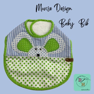 以下が含まれることがあります： 緑と白のベビーよだれかけで、マウスのデザインが施されています。よだれかけの下部は緑色の水玉模様、上部は青と白のギンガムチェック模様になっています。マウスは緑色の耳と黒いボタンの鼻をしています。よだれかけは緑色の縁取りと、"Dancing Needles"と書かれた茶色の革タグが付いています。