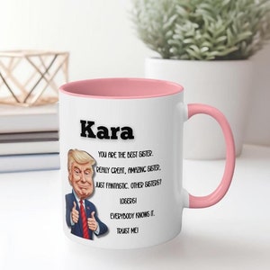 Puede incluir: Taza de cerámica blanca con interior y asa rosas. La taza presenta el nombre "Kara" en letras negras en negrita, junto con una imagen de dibujos animados de un hombre y el texto "You are the best sister. Really great, amazing sister. Just fantastic. Other sisters? Losers! Everybody knows it. Trust me!"