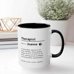 Puede incluir: Taza de cerámica blanca con borde y asa negros. La taza tiene un diseño en blanco y negro que dice "Therapist Noun (n.) Joanna" con un icono de altavoz. El texto debajo del nombre dice "Un individuo alentador, solidario y paciente, cuya guía cambia la vida de los demás para mejor, inspirando crecimiento, confianza y transformaciones positivas duraderas. Ver también: hacedor de milagros, superhéroe."