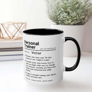 May include: White coffee mug with a black rim and handle. The mug has a black and white design with the definition of "Personal Trainer" and the word "Victor".