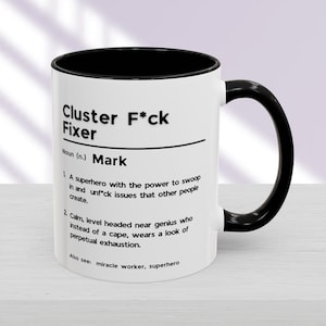 Op de afbeelding: Een witte koffiemok met een zwarte rand en handvat. De mok heeft zwarte tekst die luidt "Cluster F*ck Fixer Zelfstandig naamwoord (n.) Mark 1. Een superheld met de kracht om in te vliegen en problemen op te lossen die andere mensen veroorzaken. 2. Rustig, nuchter, bijna een genie die in plaats van een cape, een blik van voortdurende uitputting draagt. Zie ook: wonderdoener, superheld"
