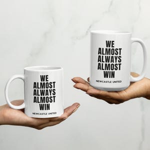 May include: Two white ceramic mugs with black text that reads "WE ALMOST ALWAYS ALMOST WIN" and "NEWCASTLE UNITED". One mug is held in each hand. The mugs have a white background and a curved handle.