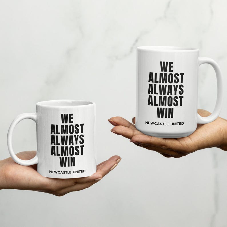 May include: Two white ceramic mugs with black text that reads "WE ALMOST ALWAYS ALMOST WIN" and "NEWCASTLE UNITED". One mug is held in each hand. The mugs have a white background and a curved handle.
