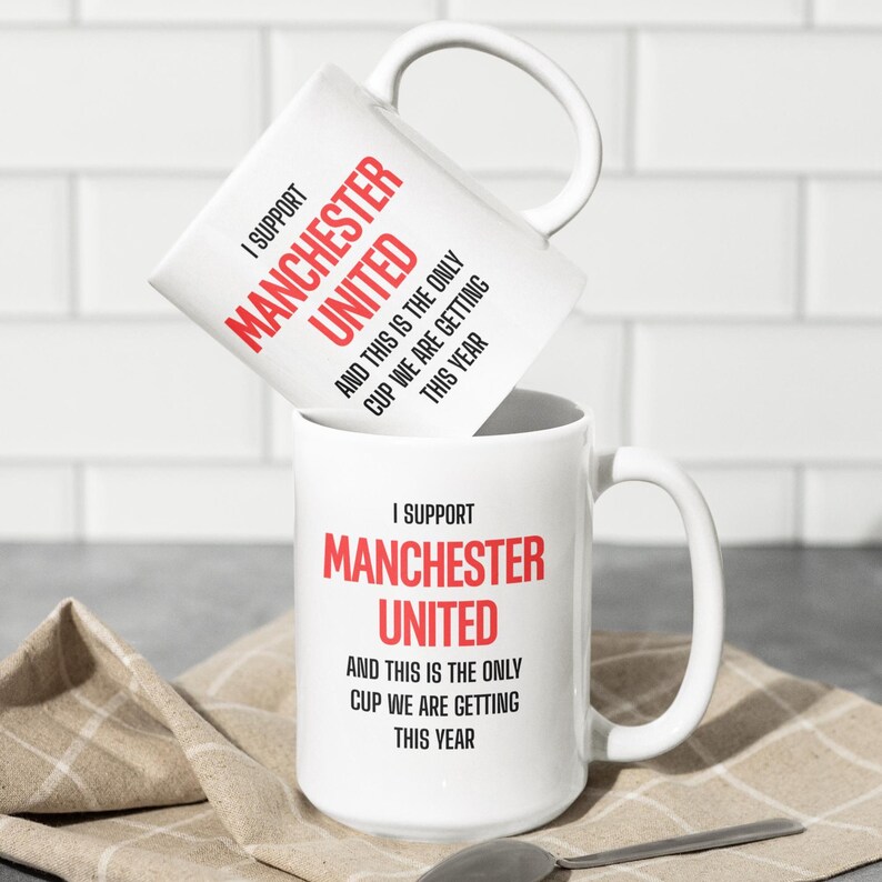 Op de afbeelding: Twee witte keramische mokken met grote handvatten. De mokken hebben de tekst "I SUPPORT MANCHESTER UNITED" in rood en zwart. Daaronder staat de zin "AND THIS IS THE ONLY CUP WE ARE GETTING THIS YEAR."