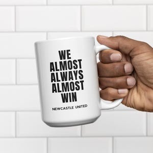 May include: White ceramic mug with the text "WE ALMOST ALWAYS ALMOST WIN" in bold black letters. The words "NEWCASTLE UNITED" are printed below the main text in a smaller font. The mug has a white handle.
