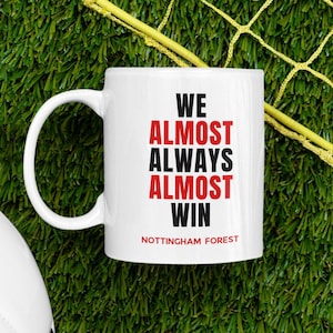Può includere: Tazza in ceramica bianca con la scritta "WE ALMOST ALWAYS ALMOST WIN" in nero e rosso, con "NOTTINGHAM FOREST" sotto. La tazza è su una superficie verde con una rete gialla.