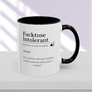 Könnte beinhalten: Weißer Keramikbecher mit schwarzem Innenraum und Henkel. Der Becher zeigt den Text "Fucktose Intolerant" mit phonetischer Aussprache und einer Definition, die lautet: "Der Zustand, andere Leute's Bullshit nicht tolerieren zu können."