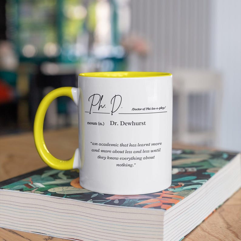 May include: White coffee mug with a yellow rim. The mug has a black inscription that reads "Ph. D. Doctor of Phi-los-o-phy/ noun (n.) Dr. Dewhurst 'an academic that has learnt more and more about less and less until they know everything about nothing.'"