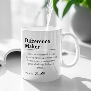 Può includere: Una tazza in ceramica bianca con testo stampato in nero che recita Difference Maker, seguita da una definizione che descrive qualcuno le cui piccole azioni creano grandi increspature, e il testo see also: Justin in basso.