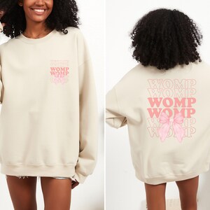 Può includere: Una felpa beige con un fiocco rosa e la parola "WOMP" ripetuta più volte sul retro.