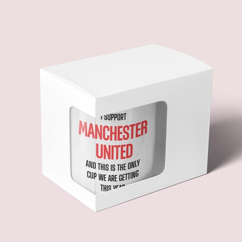 Op de afbeelding: Witte keramische mok met de tekst "I SUPPORT MANCHESTER UNITED" in rood en zwart. De mok zit in een witte doos met een uitgesneden venster. De tekst "AND THIS IS THE ONLY CUP WE ARE GETTING THIS YEAR" staat ook op de mok.