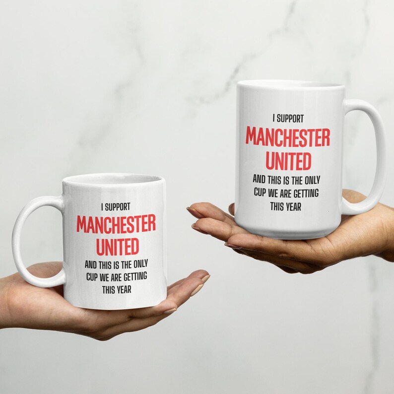 Op de afbeelding: Twee witte keramische mokken met de tekst "I SUPPORT MANCHESTER UNITED AND THIS IS THE ONLY CUP WE ARE GETTING THIS YEAR" in rood en zwart. De mokken worden voor een witte achtergrond gehouden.