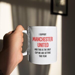 Op de afbeelding: Witte keramische mok met de tekst "I SUPPORT MANCHESTER UNITED AND THIS IS THE ONLY CUP WE ARE GETTING THIS YEAR" in rood en zwart. De mok wordt door een hand vastgehouden.