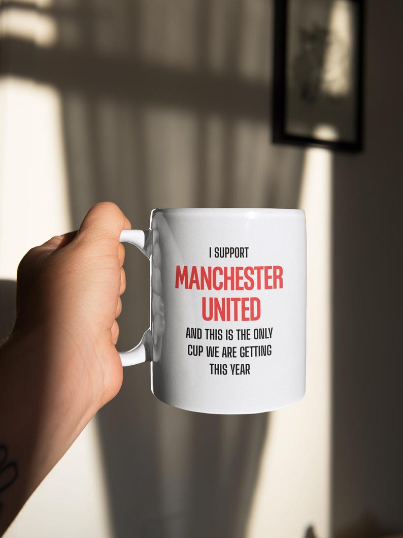 Op de afbeelding: Witte keramische mok met de tekst "I SUPPORT MANCHESTER UNITED AND THIS IS THE ONLY CUP WE ARE GETTING THIS YEAR" in rood en zwart. De mok wordt door een hand vastgehouden.