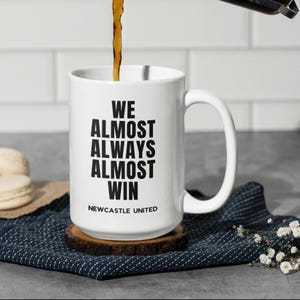 May include: White ceramic mug with the text "WE ALMOST ALWAYS ALMOST WIN" in black, with "NEWCASTLE UNITED" below. The mug sits on a wooden coaster, with coffee being poured into it. The mug has a large handle.