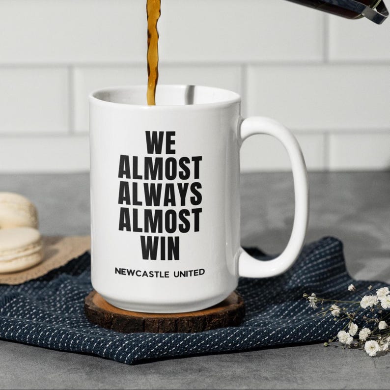 May include: White ceramic mug with the text "WE ALMOST ALWAYS ALMOST WIN" in black, with "NEWCASTLE UNITED" below. The mug sits on a wooden coaster, with coffee being poured into it. The mug has a large handle.