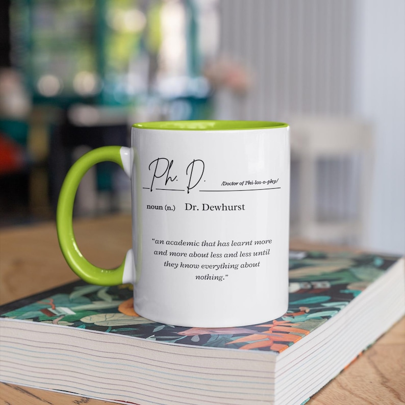 May include: White coffee mug with a green rim. The mug has black text that reads "Ph. D. Doctor of Phi-los-o-phy/ noun (n.) Dr. Dewhurst 'an academic that has learnt more and more about less and less until they know everything about nothing.'"