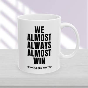 May include: White ceramic mug with the text "WE ALMOST ALWAYS ALMOST WIN" in black, with "NEWCASTLE UNITED" below. The mug has a curved handle and is on a white surface.