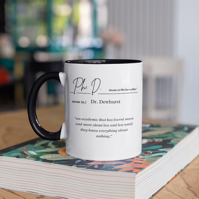 May include: White coffee mug with a black handle and black rim. The mug has a black inscription that reads "Ph. D. Doctor of Phi-los-o-phy/ noun (n.) Dr. Dewhurst 'an academic that has learnt more and more about less and less until they know everything about nothing.'"