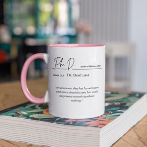 May include: White ceramic mug with a pink rim and handle. The mug has a black inscription that reads "Ph.D. Doctor of Phi-los-o-phy/ noun (n.) Dr. Dewhurst 'an academic that has learnt more and more about less and less until they know everything about nothing.'"