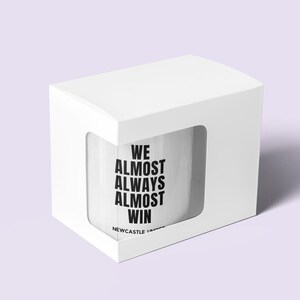 May include: White ceramic mug with the text "WE ALMOST ALWAYS ALMOST WIN" in black, inside a white box with a window. The mug also has the text "NEWCASTLE UNITED" in black.