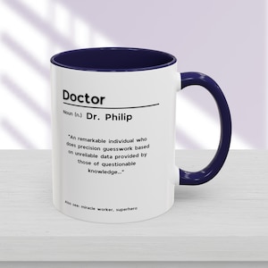 Puede incluir: Taza de cerámica blanca con interior y asa azul marino. La taza presenta la palabra "Doctor" sobre una definición que incluye el nombre "Dr. Philip" y una descripción humorística. También se incluye el texto "Also see: miracle worker, superhero".