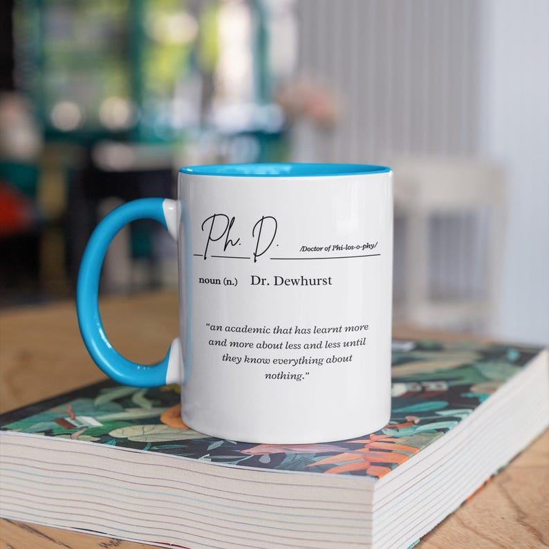 May include: White ceramic mug with a blue rim and handle. The mug has black text that reads "Ph. D. Doctor of Phi-los-o-phy/ noun (n.) Dr. Dewhurst 'an academic that has learnt more and more about less and less until they know everything about nothing.'"