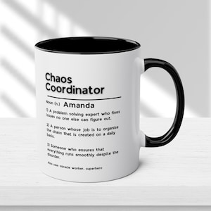 Op de afbeelding: Witte keramische mok met een zwarte rand en handvat. De mok heeft een zwart-witte afbeelding met de tekst "Chaos Coordinator" en een definitie van de term, inclusief de naam "Amanda".
