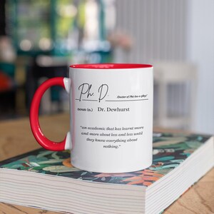 May include: White coffee mug with a red rim and handle. The mug has black text that reads "Ph. D. Doctor of Phi-los-o-phy/ noun (n.) Dr. Dewhurst 'an academic that has learnt more and more about less and less until they know everything about nothing.'"