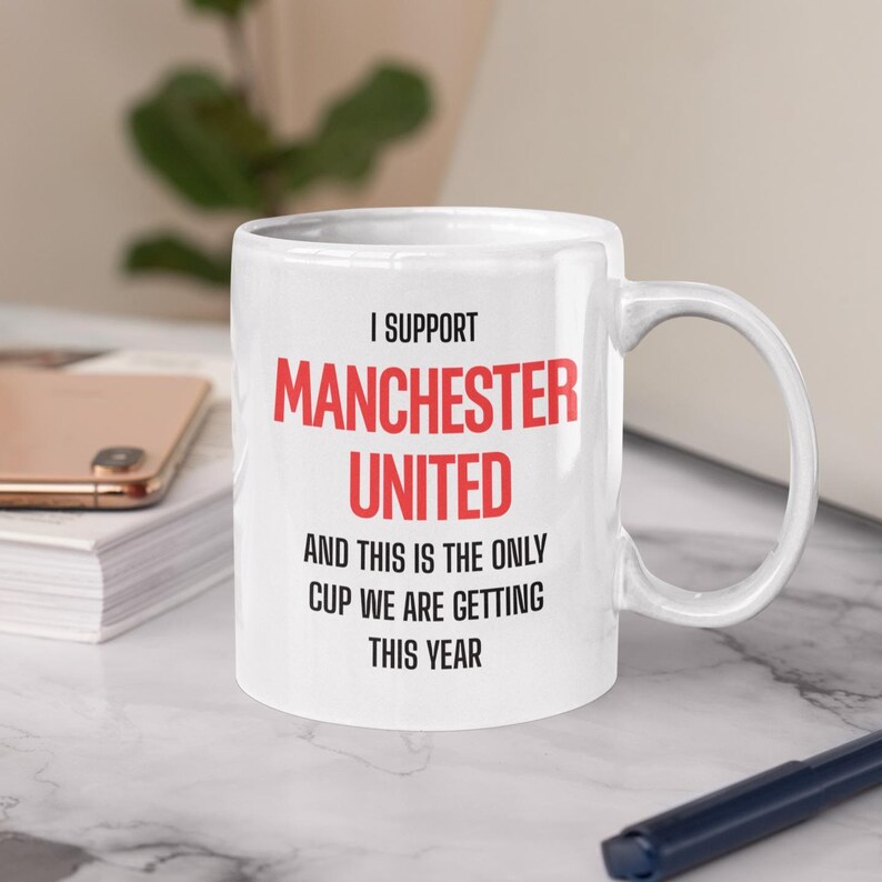 Op de afbeelding: Witte keramische mok met de tekst "I SUPPORT MANCHESTER UNITED" in rood en zwart. De mok zegt ook "AND THIS IS THE ONLY CUP WE ARE GETTING THIS YEAR". De mok heeft een gebogen handvat.