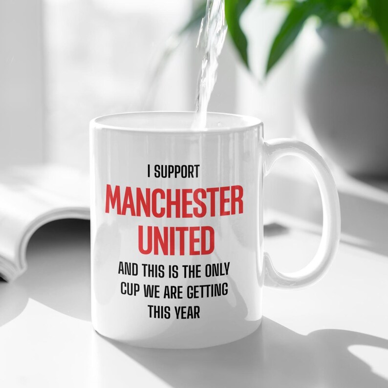 Op de afbeelding: Witte keramische mok met de tekst "I SUPPORT MANCHESTER UNITED" in zwart en rood. Daaronder staat "AND THIS IS THE ONLY CUP WE ARE GETTING THIS YEAR". De mok heeft een gebogen oor en wordt gevuld met water.