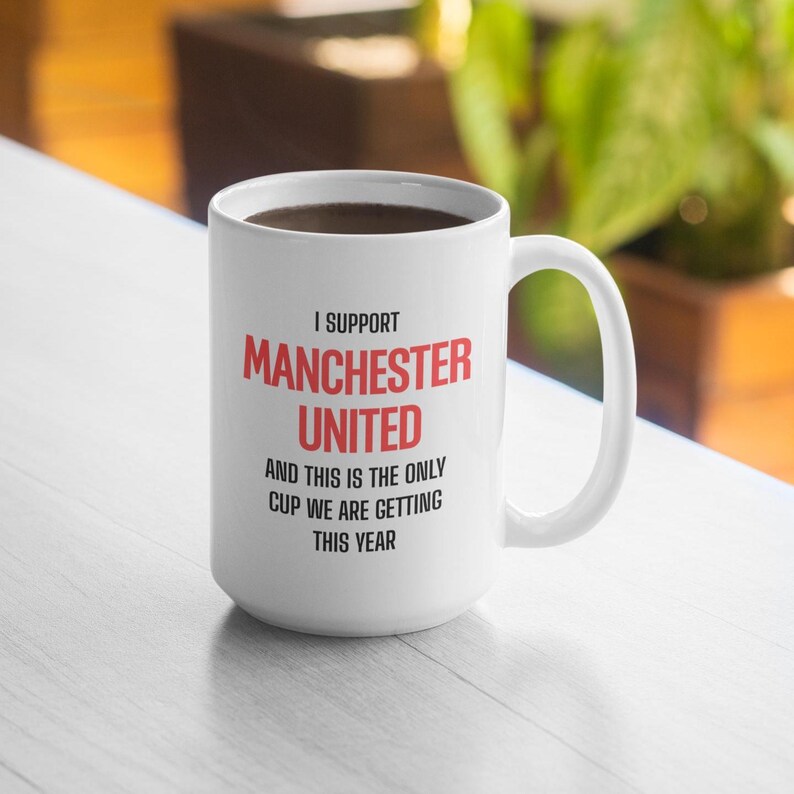 Op de afbeelding: Witte keramische mok met de tekst "I SUPPORT MANCHESTER UNITED" in rood en zwart. De mok heeft ook de tekst "AND THIS IS THE ONLY CUP WE ARE GETTING THIS YEAR". De mok is gevuld met een donkere vloeistof.