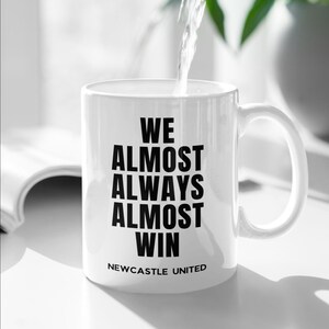 May include: White ceramic mug with the text "WE ALMOST ALWAYS ALMOST WIN" in black, with "NEWCASTLE UNITED" below. Water is being poured into the mug. The mug has a curved handle and is on a white surface.