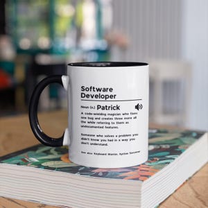 Pu&ograve; includere: Una tazza in ceramica bianca con manico e bordo neri. La tazza presenta una grafica in bianco e nero con il testo "Software Developer" e la definizione "Sostantivo (n.) Patrick Un mago del codice che risolve un bug e ne crea altri tre, mentre li definisce come funzionalit&agrave; non documentate. Qualcuno che risolve un problema che non sapevi di avere in un modo che non capisci. Vedi anche: Guerriero della tastiera. Stregone della sintassi"