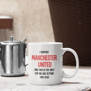 Op de afbeelding: Witte keramische mok met de tekst "I SUPPORT MANCHESTER UNITED AND THIS IS THE ONLY CUP WE ARE GETTING THIS YEAR" in zwart en rood. De mok heeft een gebogen handvat en staat op een wit oppervlak.