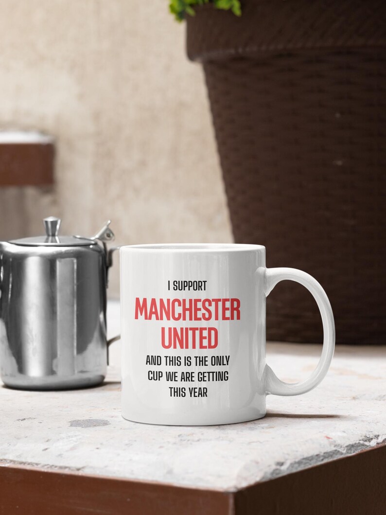 Op de afbeelding: Witte keramische mok met de tekst "I SUPPORT MANCHESTER UNITED AND THIS IS THE ONLY CUP WE ARE GETTING THIS YEAR" in zwart en rood. De mok heeft een gebogen handvat en staat op een wit oppervlak.