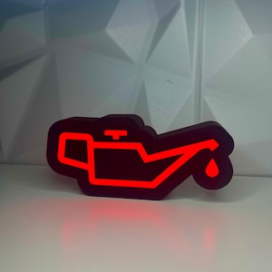 May include: A black and red oil warning light. The symbol is a stylized oil can with a red outline and a red drop of oil. The light is illuminated, indicating a potential issue with the vehicle's oil system.