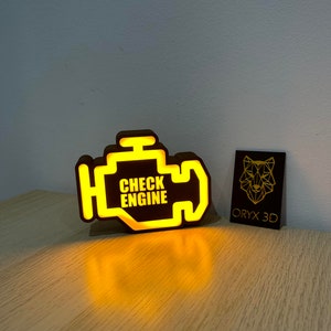 Könnte beinhalten: Ein gelbes, beleuchtetes Check-Engine-Lichtzeichen mit dem Text "CHECK ENGINE" in schwarzer Schrift. Das Schild ist aus dunkelbraunem Material und steht auf einer hellbraunen Holzoberfläche. Eine Visitenkarte mit dem Text "ORYX 3D" ist im Hintergrund sichtbar.