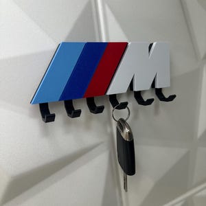 Op de afbeelding: Een aan de muur gemonteerde sleutelhouder met het BMW M-logo in blauw, donkerblauw, rood en wit. Het heeft vijf zwarte haken, met een autosleutel die aan &eacute;&eacute;n ervan hangt. De sleutelhouder is gemonteerd op een witte, getextureerde muur.