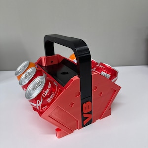 May include: Red and black 3D printed can holder with a handle. The holder has the text "V8" printed on the side. The holder is filled with six aluminum cans.