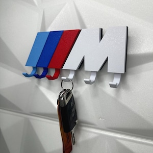 Peut inclure: Un porte-clés blanc et noir en forme de lettre "M" avec des rayures bleues, rouges et blanches. Le porte-clés comporte quatre crochets et est monté sur un mur blanc texturé. Un jeu de clés est suspendu à l'un des crochets.