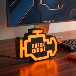 May include: A black, engine-shaped desk lamp with a glowing orange border and the text "CHECK ENGINE" illuminated in the center. The lamp features a matte black finish and a bright orange light that outlines the iconic automotive symbol.