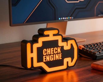 Op de afbeelding: Een zwarte, motorvormige bureaulamp met een gloeiende oranje rand en de tekst "CHECK ENGINE" verlicht in het midden. De lamp heeft een matzwarte afwerking en een fel oranje licht dat het iconische autosymbool omlijnt.