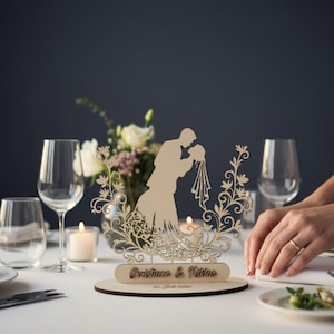 Könnte beinhalten: Ein hölzerner Tortenaufsatz für eine Hochzeit mit einer Silhouette eines sich küssenden Paares, umgeben von floralen und Rankenmustern. Die Basis enthält die Namen "Christiane & Niklas" und das Hochzeitsdatum. Der Aufsatz steht auf einer runden Holzbasis.
