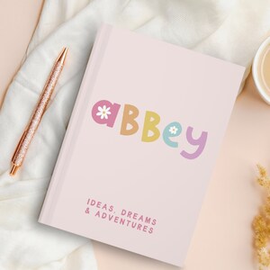 Może przedstawiać: R&oacute;żowy notes z imieniem "Abbey" w kolorowych literach z kwiatowym wzorem. Tekst "Pomysły, marzenia i przygody" jest wydrukowany pod imieniem.
