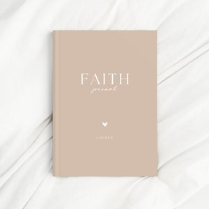 May include: A beige journal with the words "FAITH journal" in white script. A small white heart and the name "LAUREN" are below the title. The journal is closed and resting on a white, textured surface.