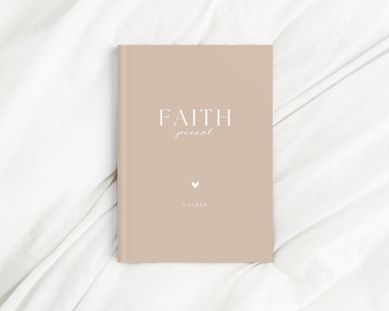 May include: A beige journal with the words "FAITH journal" in white script. A small white heart and the name "LAUREN" are below the title. The journal is closed and resting on a white, textured surface.