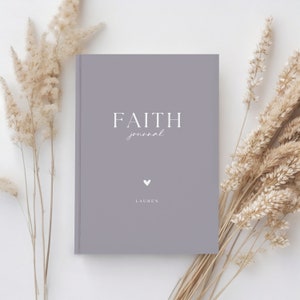 May include: A gray journal with the words "FAITH journal" in white script and a small heart symbol. The name "LAUREN" is printed below the heart. Dried beige plants are arranged around the journal on a white surface.