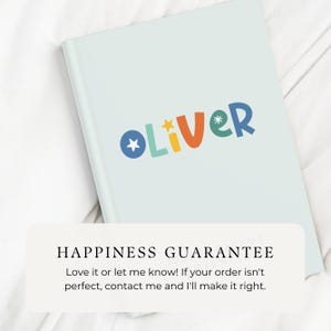 May include: A light blue notebook with the name "OLIVER" written in colorful letters. The notebook has a happiness guarantee message printed on the bottom.